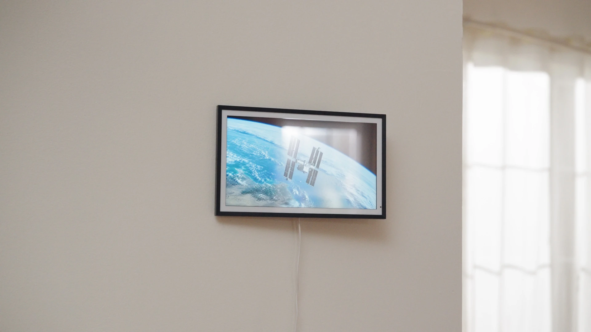 Photograph of a black LCD frame showing Earth and a space station, with window reflections and a power cable.